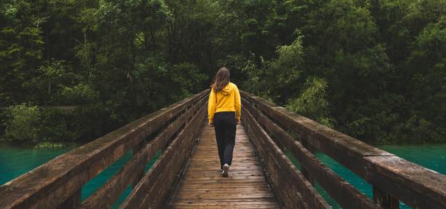 Woman in a yellow jacket walking across a bridge surrounded by a forest